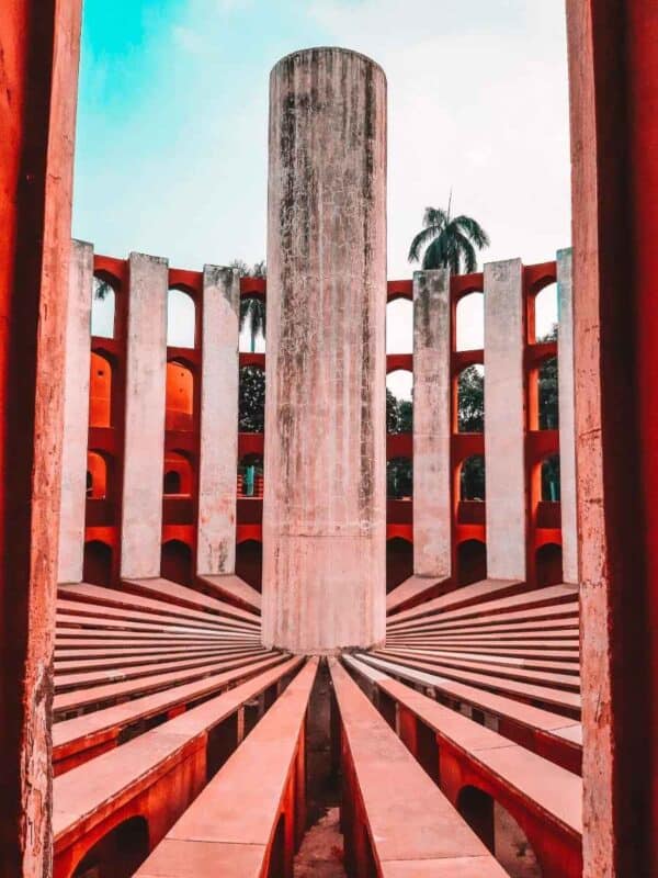 close-up of one of the astronomical instruments at jantar mantar in delhi, showing precise geometric design with symmetrical arches, red-and-white stone structure, and a tall cylindrical pillar at the center against a partly cloudy sky