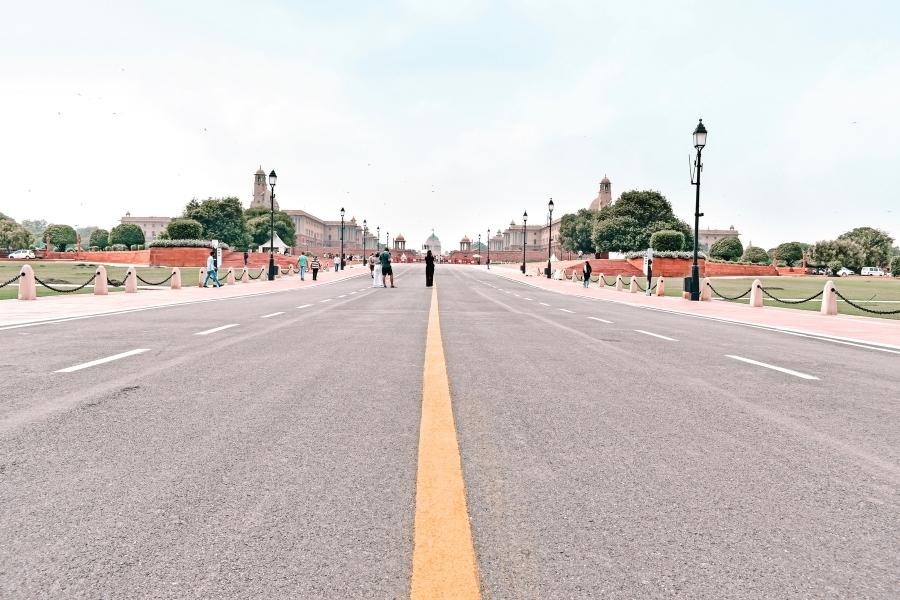 a wide ceremonial boulevard, kartavya path, formerly known as rajpath, leading to rashtrapati bhavan in new delhi, flanked by government buildings and lawns, with people walking casually along the center and sidewalks under a pale blue sky