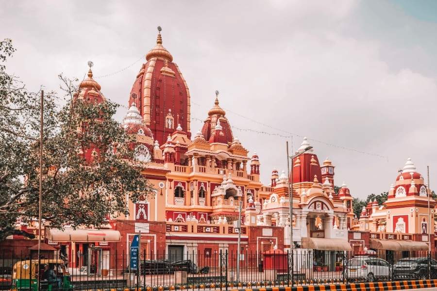 lakshmi narayan mandir (birla mandir) in delhi with its red and cream towers standing tall under a cloudy sky, surrounded by trees, auto-rickshaws, and cars on a busy street