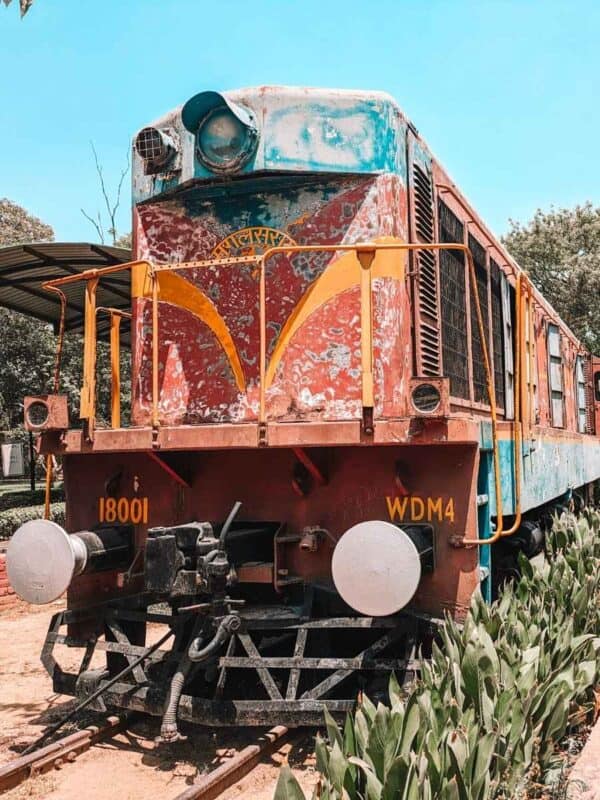 old rusted locomotive on display at the national rail museum in delhi, surrounded by greenery and a red-tiled walkway under a clear blue sky
