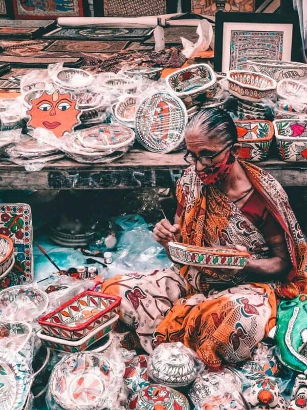 a woman in a printed saree hand-painting madhubani artwork on a bowl, surrounded by stacks of vibrantly painted traditional items at a crafts stall