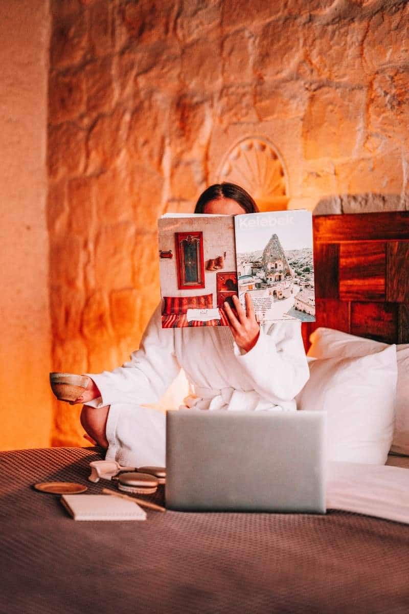 person reading a travel magazine on a bed with laptop and coffee