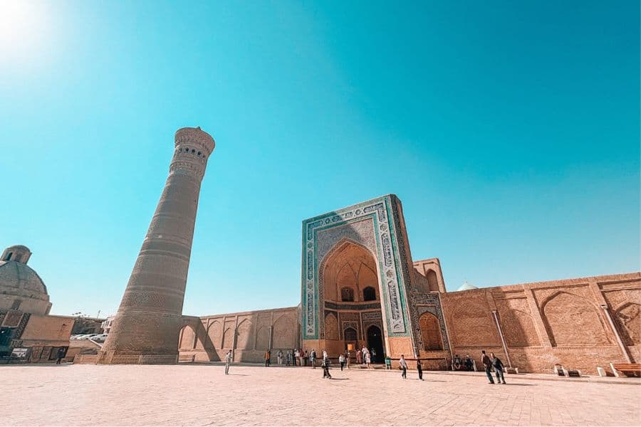 poi kalyan complex in bukhara with people walking near the minaret and turquoise-tiled madrasa under a clear sky