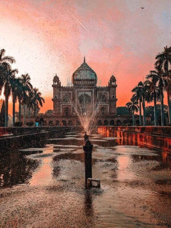 low-angle view of safdarjung tomb in delhi captured at sunset with water droplets frozen mid-air from a central fountain, flanked by palm trees and a warm sky streaked with light