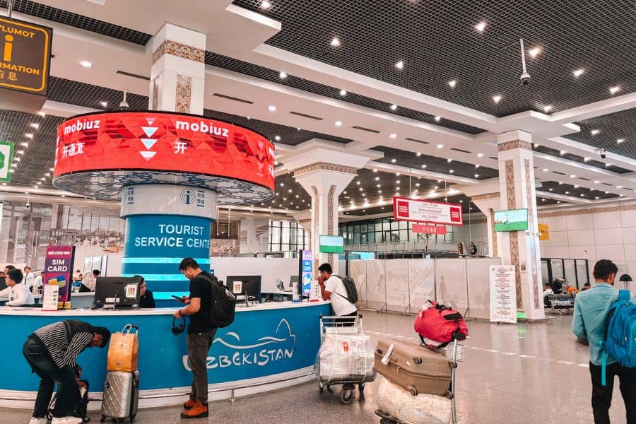 tourist service center at tashkent international airport with sim card counters, information desk, travelers with luggage, and clear signage inside a bright terminal hall