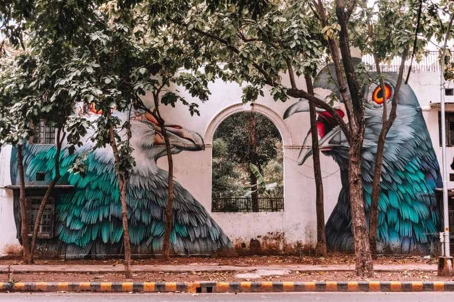 vibrant mural of giant pigeons painted across a white building wall in lodhi art district, delhi’s first open-air public art gallery, partially hidden behind trees along a quiet roadside