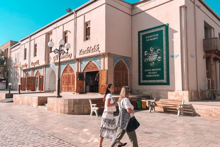 zardozlik uy muzeyi in bukhara, house museum of gold embroidery with arched wooden doors, green museum signboard on the wall, and two women walking across the stone-paved courtyard under a clear blue sky
