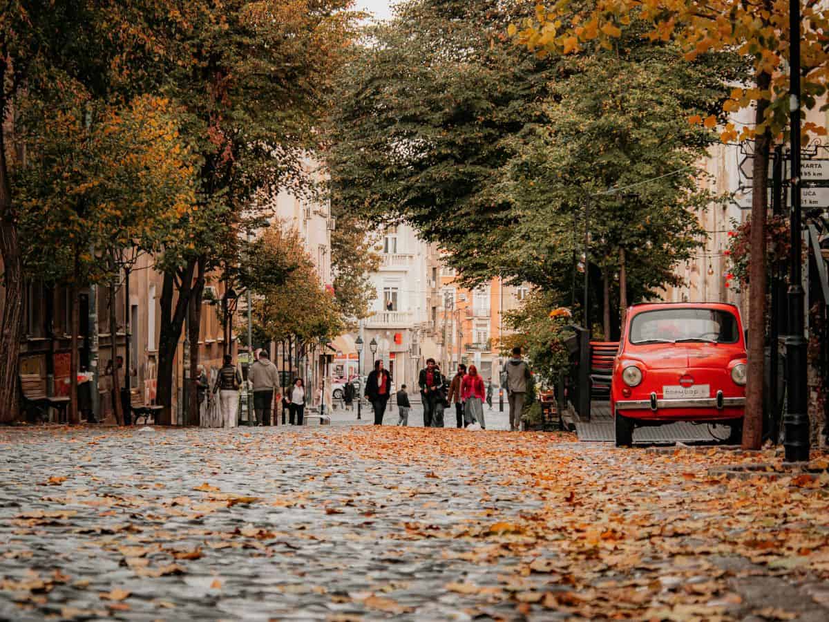 Autumn street scene in Europe with fallen leaves covering cobblestones, red vintage car parked under tree-lined boulevard, and people walking leisurely during shoulder season travel