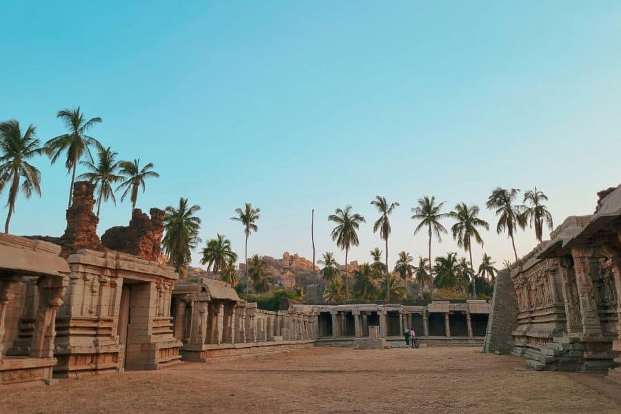 Ancient temple ruins at Hampi, Karnataka, with carved stone colonnades on either side of a wide open courtyard, tall palm trees rising against a clear blue sky, and the characteristic boulder-strewn hills of the Vijayanagara landscape in the background.
