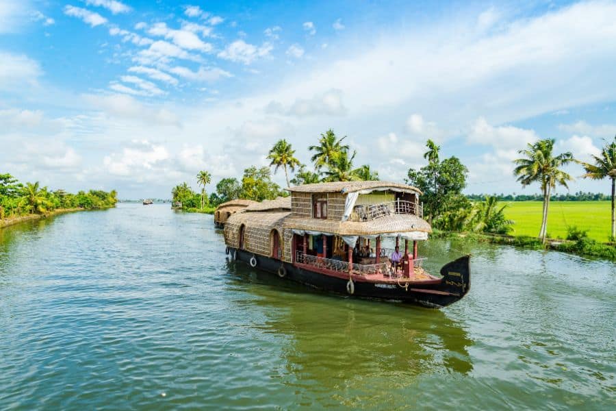 A traditional Kerala kettuvallam houseboat drifting along a calm backwater canal in Alleppey, surrounded by coconut palms and bright green rice paddies under a partly cloudy blue sky.