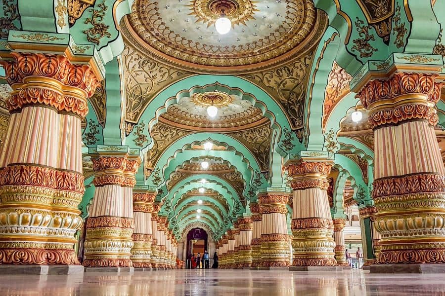 The ornate interior of Mysore Palace showing grand arched corridors, intricately carved columns in terracotta and gold, and a decorative domed ceiling painted in mint green and gold, stretching into the distance under warm pendant lights.