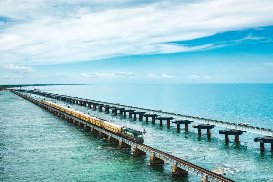 A yellow and green Indian Railways train crossing the Pamban Bridge over the turquoise waters of the Palk Strait, connecting mainland India to Rameshwaram island under a wide open sky.
