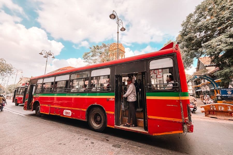 A red BEST bus on the streets of Colaba, South Mumbai, with a commuter standing at the open door, Mumbai's iconic public bus system