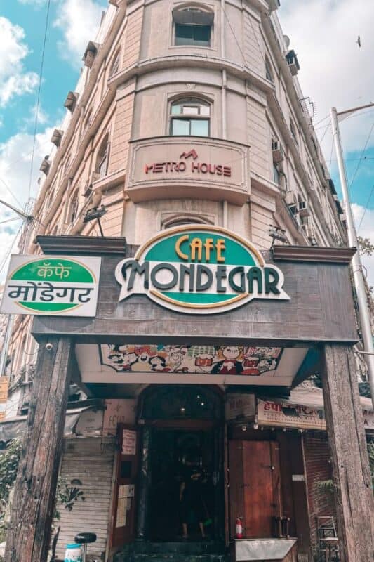 Exterior view of Cafe Mondegar at Metro House on Colaba Causeway, with the cafe's classic Art Deco signage in English and Marathi, and Mario Miranda murals visible above the entrance