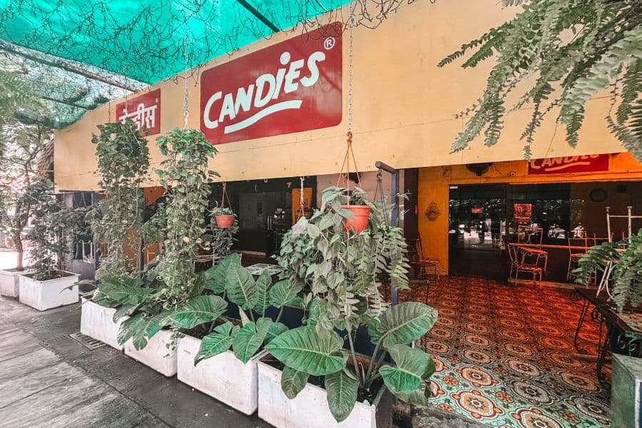 The exterior of Candies, an iconic Bandra cafe in Mumbai, with hanging ferns, patterned floor tiles, and the cafe's signature red-and-cream signage in English and Marathi
