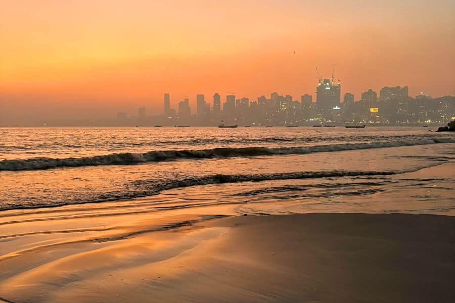 Sunset at Girgaon Chowpatty Beach in Mumbai, with traditional Koli fishing boats anchored in the calm sea and the city skyline silhouetted against an amber sky