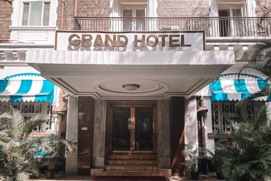 The Art Deco entrance of Grand Hotel in Ballard Estate, South Mumbai, featuring the heritage hotel's gold "Grand Hotel" signage, blue-and-white striped awnings, and original brass doors