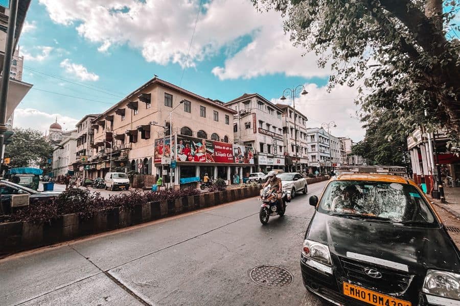 Street view of the iconic Leopold Cafe on Colaba Causeway, Mumbai, the Parsi-Irani institution running since the 1870s, made famous worldwide by Gregory David Roberts' novel Shantaram, with a kaali-peeli taxi and the Taj Mahal Palace Hotel dome glimpsed in the background