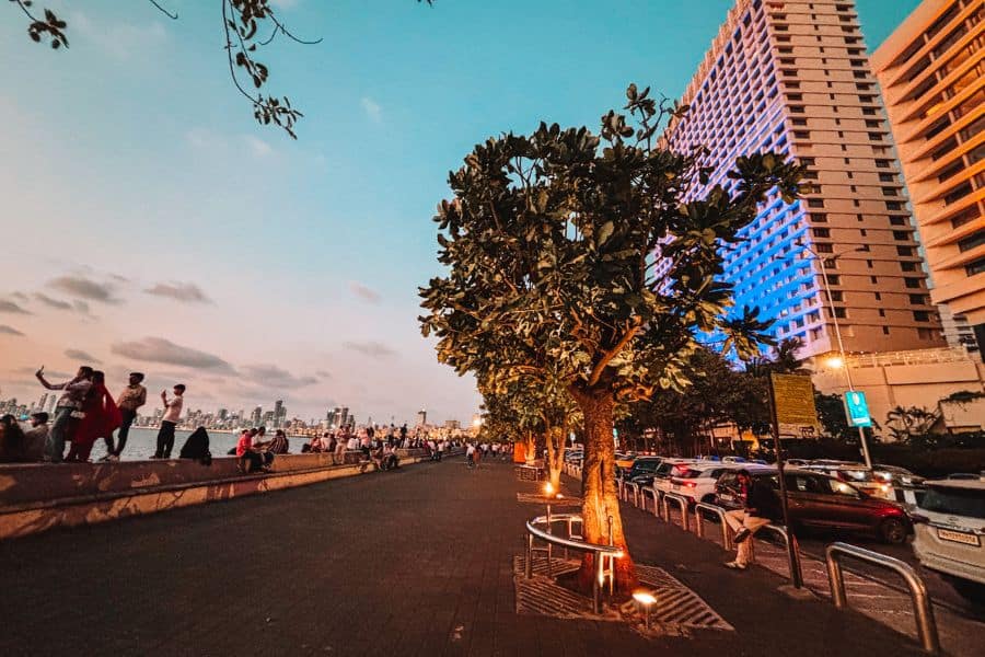 The Marine Drive promenade at dusk, with the curving walkway, lit trees, and South Mumbai skyline at blue hour, including the Air India and Express Towers buildings