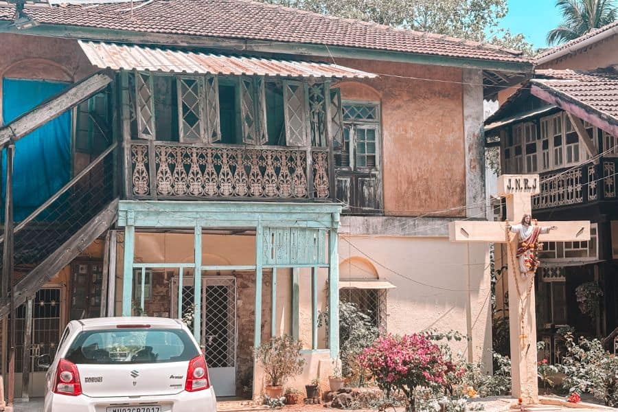 A heritage East Indian Catholic cottage in Ranwar Village, Bandra West, Mumbai, with a wayside cross dating to 1866, ornate wooden balcony railings, and sloped tile roof